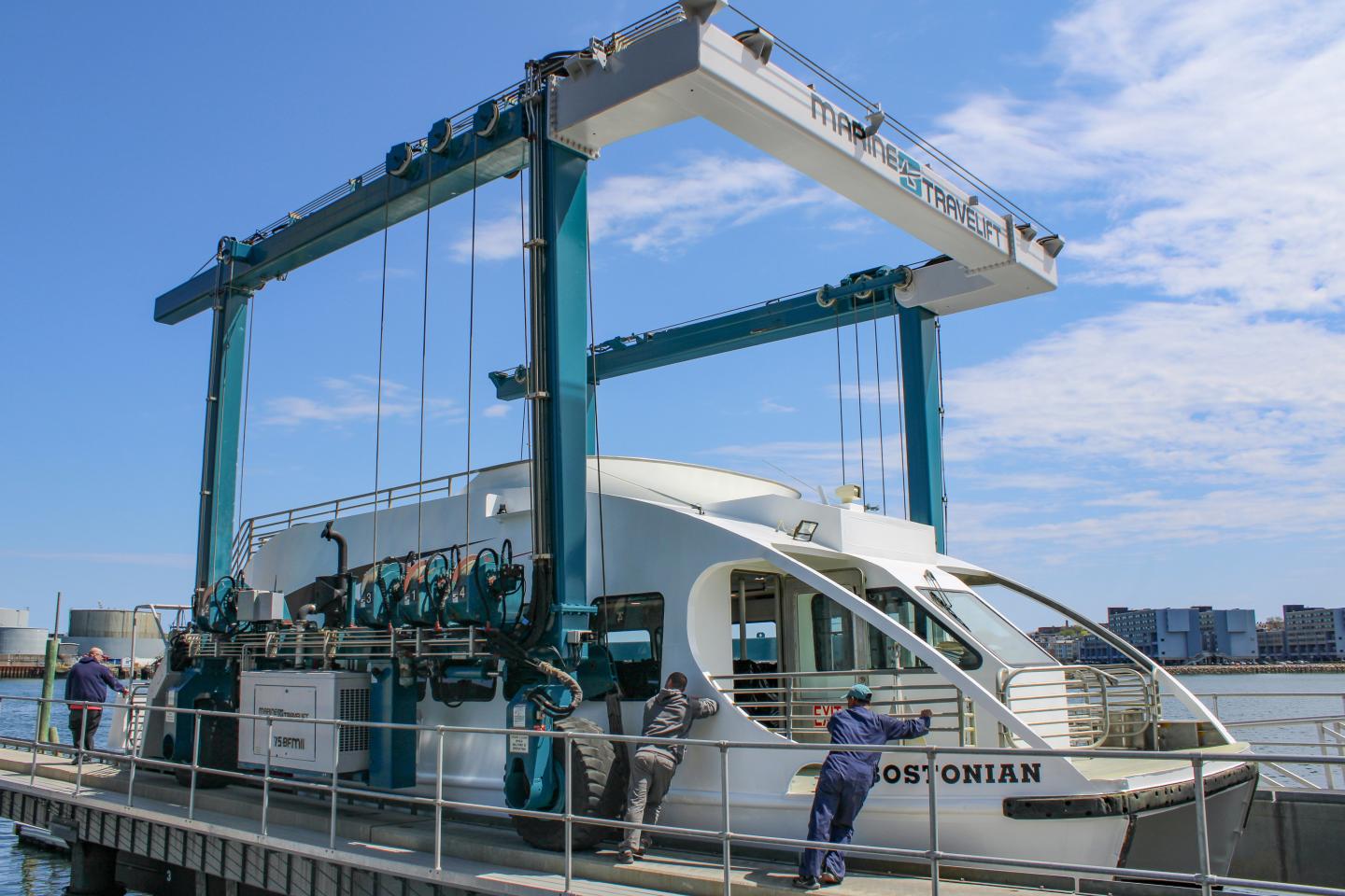 Boat lifted by large blue crane, workers nearby, under blue sky.