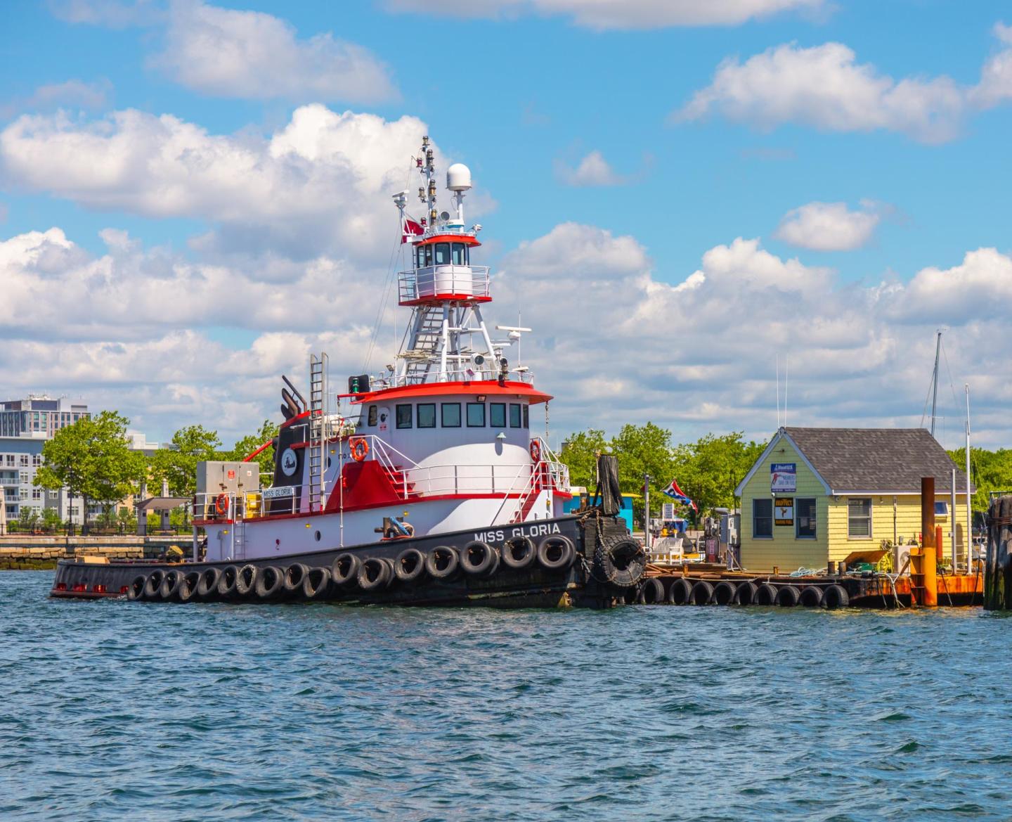 Red and white tugboat docked near a small yellow building on a sunny day.