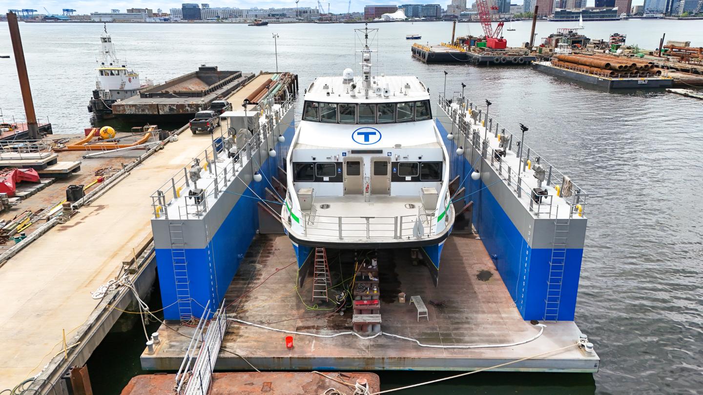 A ferry docked in a shipyard, with blue accents and a city skyline in the background.